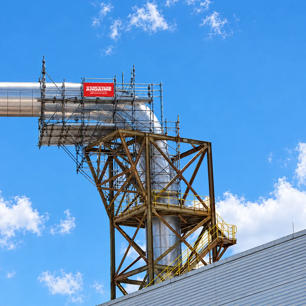 Andaime montado ao redor de uma tubulação industrial, com a placa da Andaime, sob céu azul